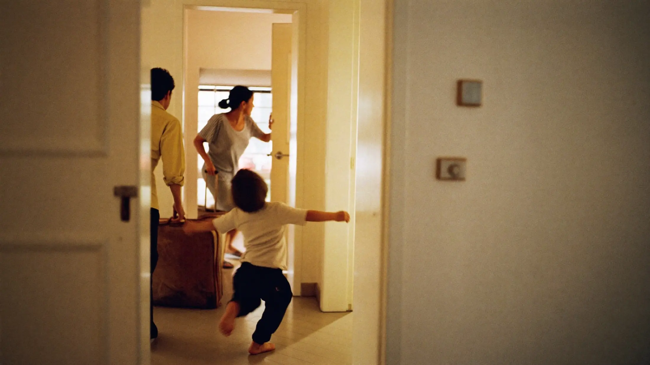 Un niño corre con los brazos abiertos hacia la puerta abierta de casa, con una maleta recién llegada en el recibidor